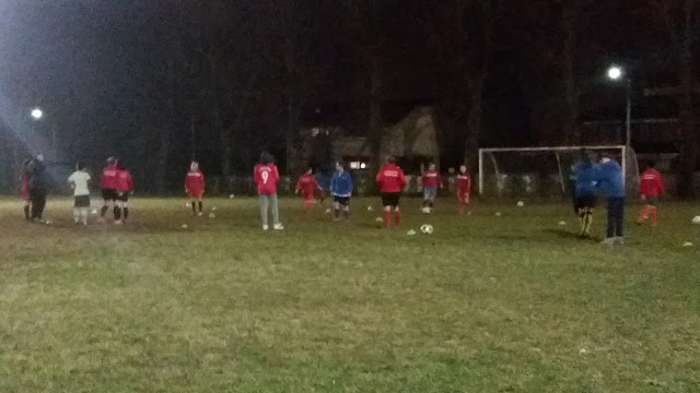 Allenamento serale di calcio femminile su un campo in erba sotto i riflettori, con giocatrici in maglia rossa e pantaloncini scuri, atmosfera intensa e sportiva