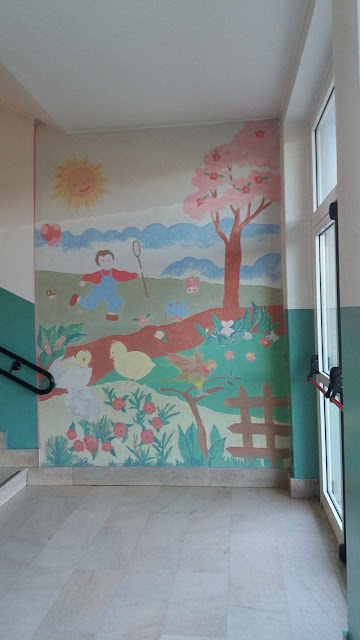 Murale dipinto su una parete con figure di bambini, papere, un albero, il sole e fiori in tonalità pastello, atmosfera allegra e comunitaria